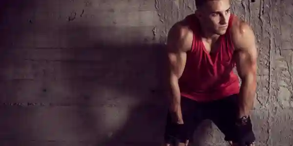 A man in red shirt and black shorts leaning against wall.